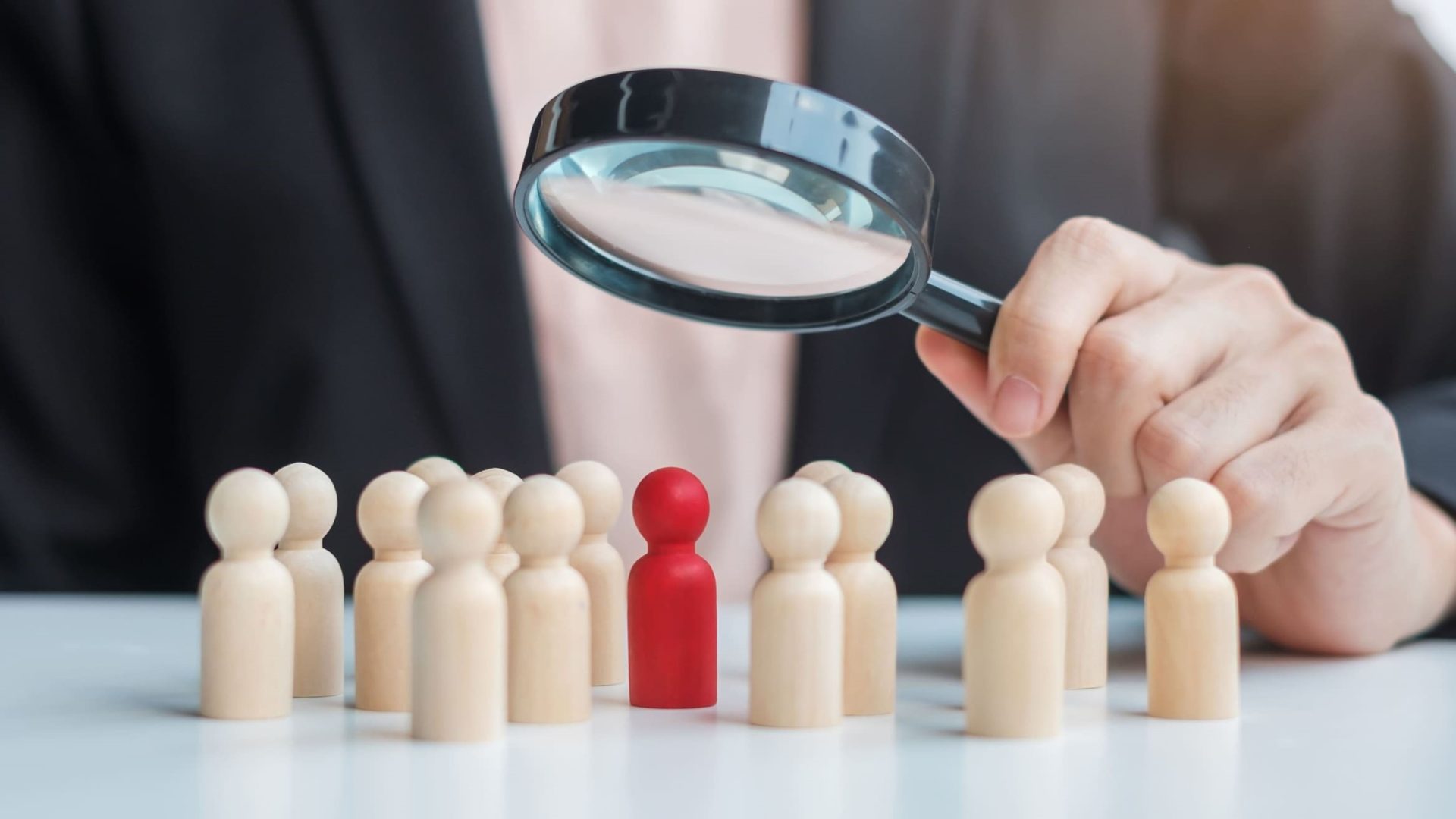 Mano de auditor sosteniendo una lupa sobre una figura roja destacada entre un grupo de figuras de madera, simbolizando el escrutinio y la revisión en procesos de auditoría voluntaria y obligatoria.