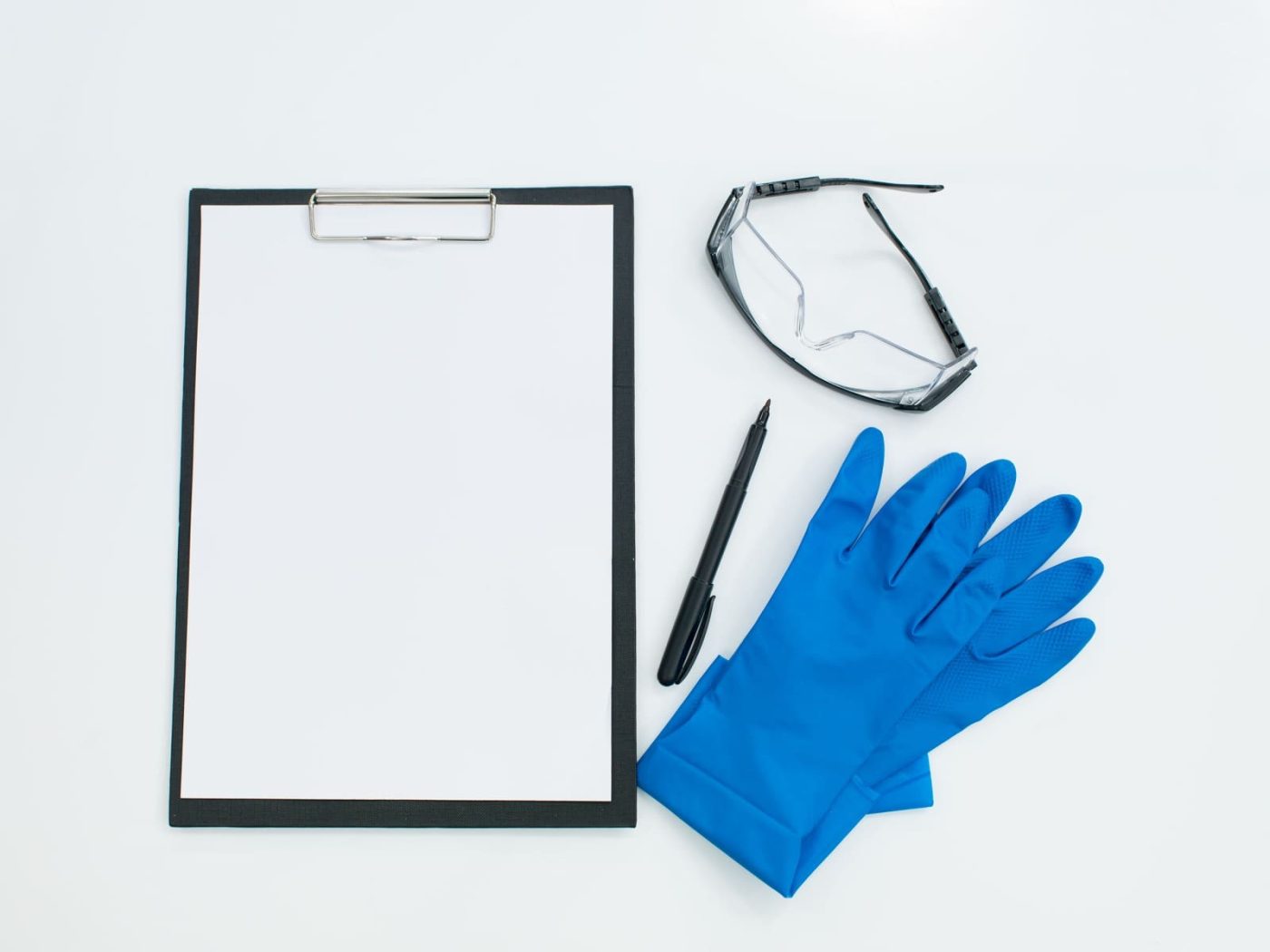 Portapapeles, guantes de protección y gafas de seguridad sobre fondo blanco, representando la formación en prevención de riesgos laborales para empleadas de hogar.