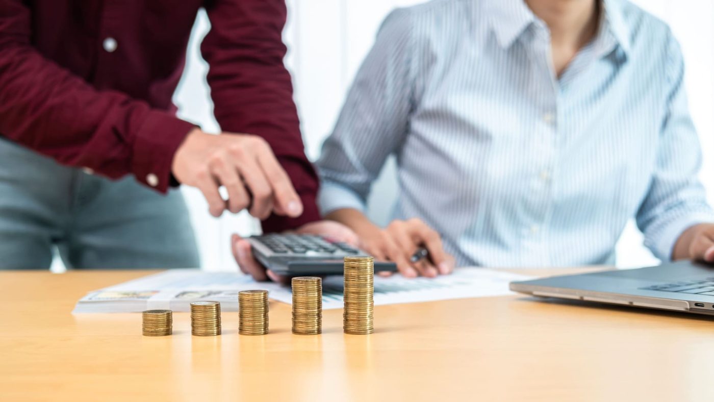 Asesor fiscal y cliente revisando cálculos junto a columnas de monedas sobre una mesa de trabajo.